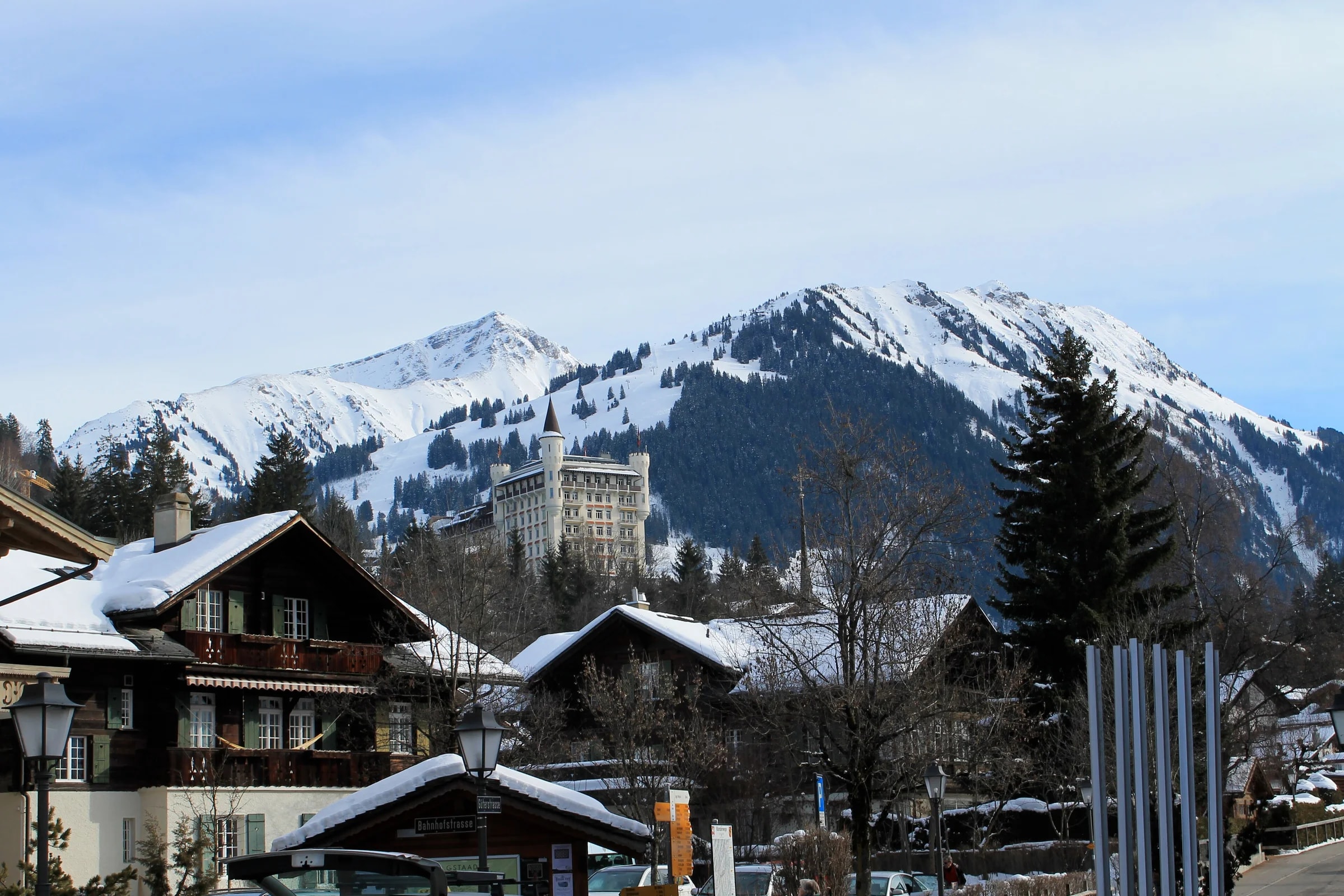 Verschneite Chalets in Gstaad, dahinter ein großes Hotel mit Türmen auf dem Hügel und die Berge.