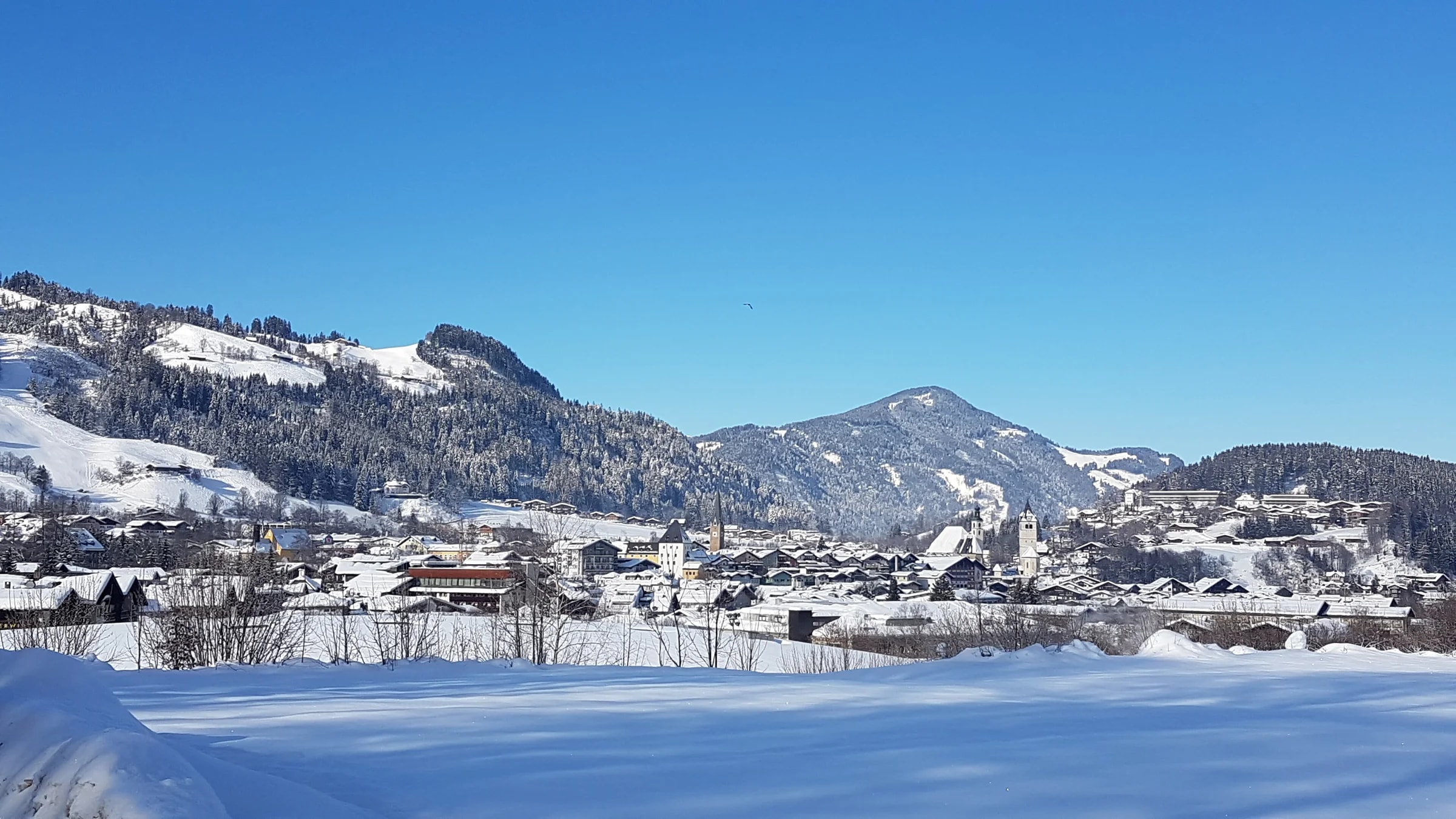 Kitzbühel im Schnee, mit seinen Kirchtürmen am Fuß bewaldeter, verschneiter Berge unter blauem Himmel.