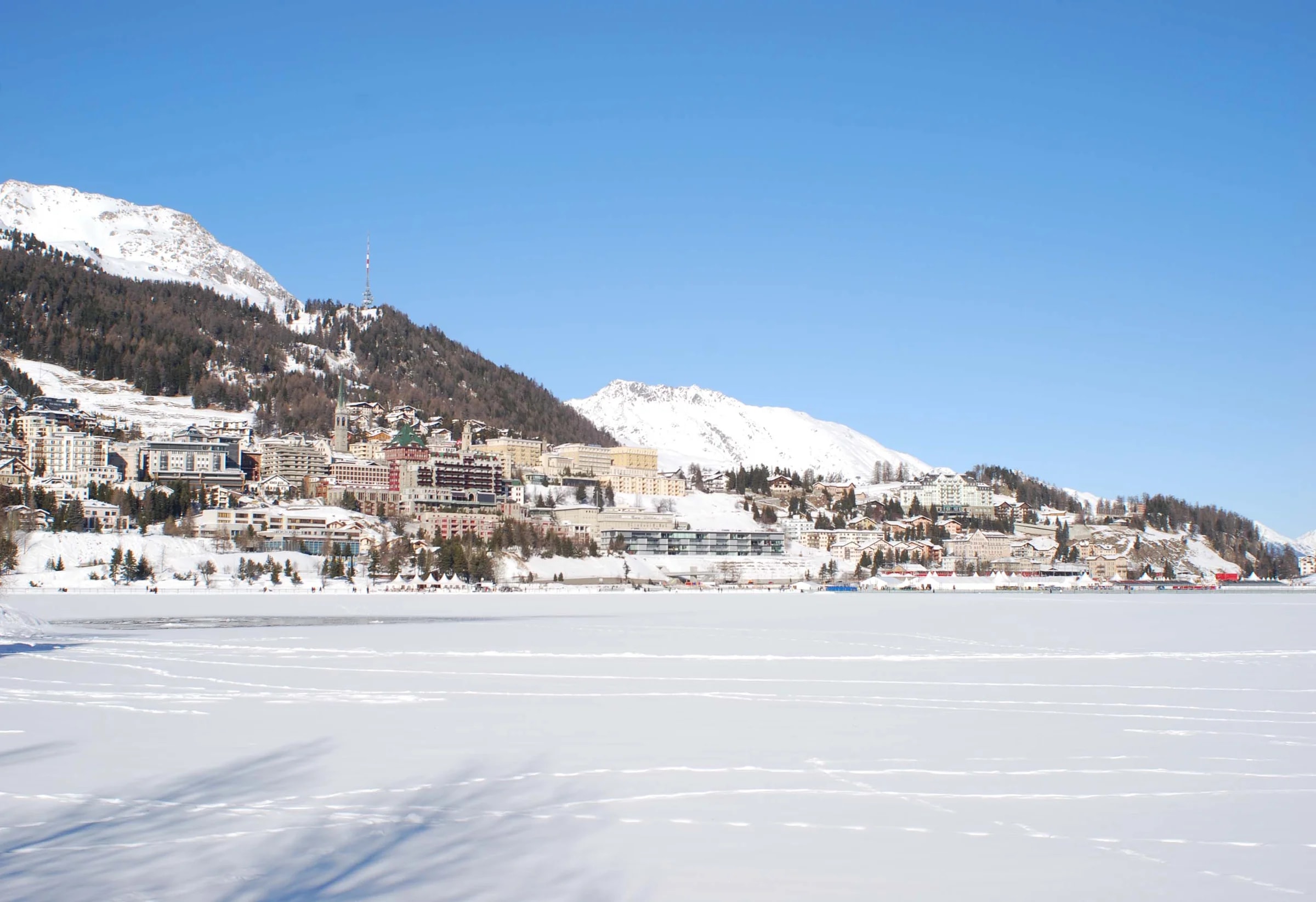 St. Moritz vom zugefrorenen, verschneiten See aus, der Ort am Hang unter den Gipfeln und blauem Himmel.
