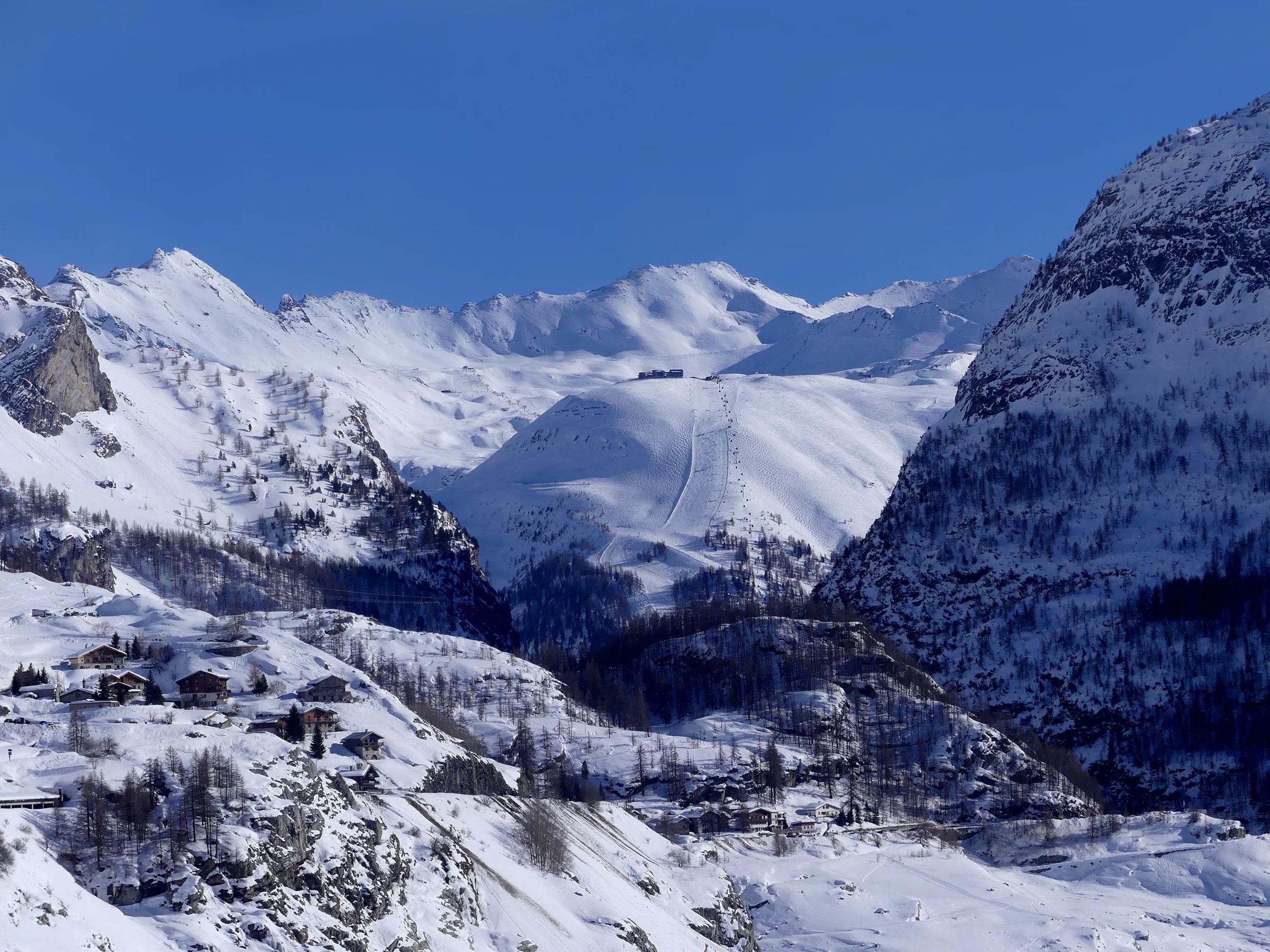 Das verschneite Skigebiet von Val d'Isère unter blauem Himmel, mit Chalets und Lärchen im Vordergrund.