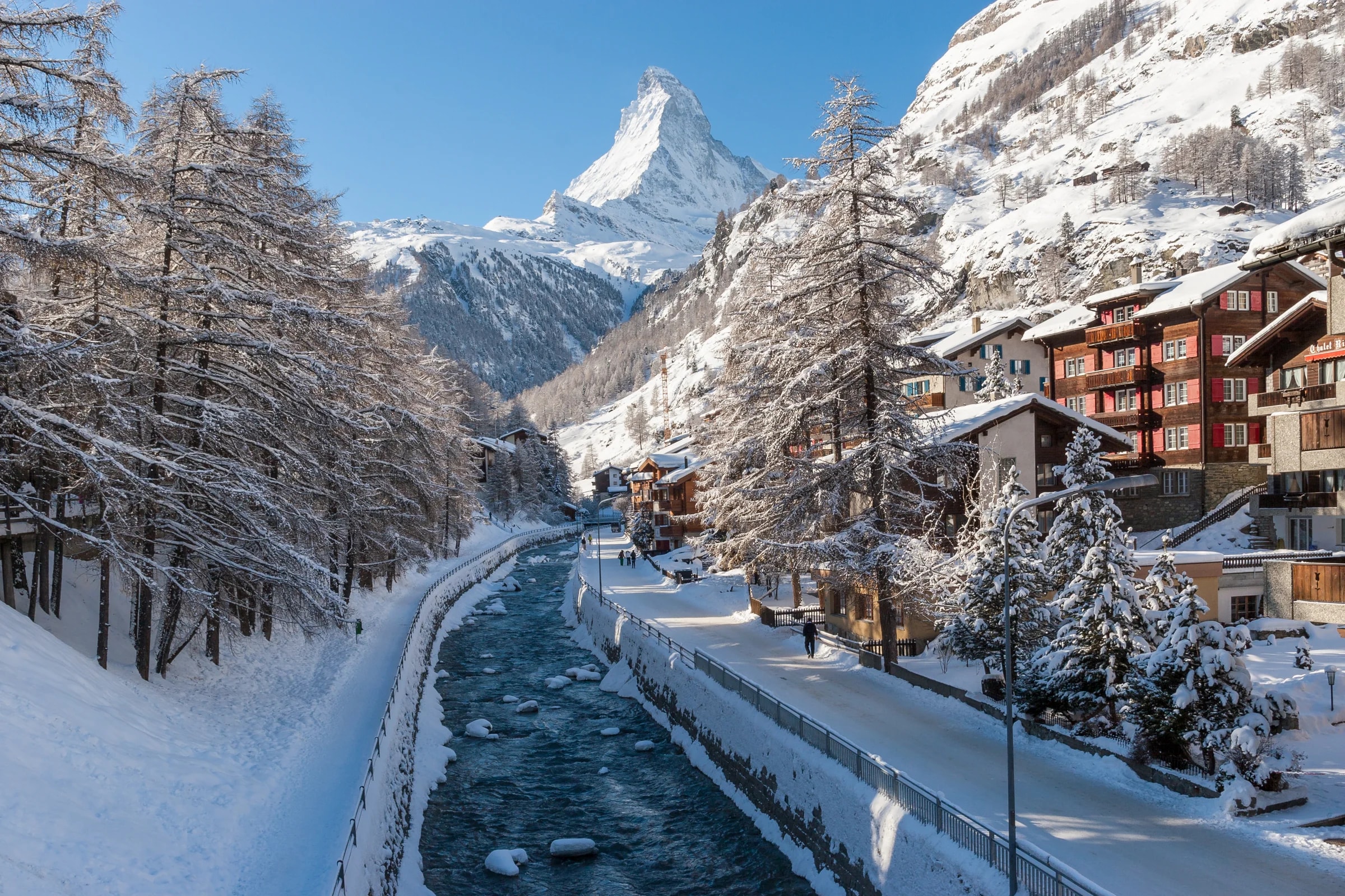 Die Vispa fließt durch das verschneite Zermatt zwischen Holzchalets und Lärchen, dahinter das Matterhorn unter blauem Himmel.