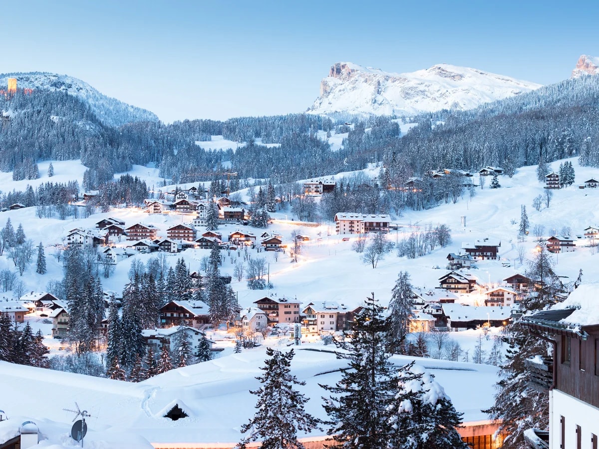 Verschneite Chalets im Weiler Ronco in Cortina d'Ampezzo bei Tagesanbruch, dahinter Gipfel der Dolomiten.