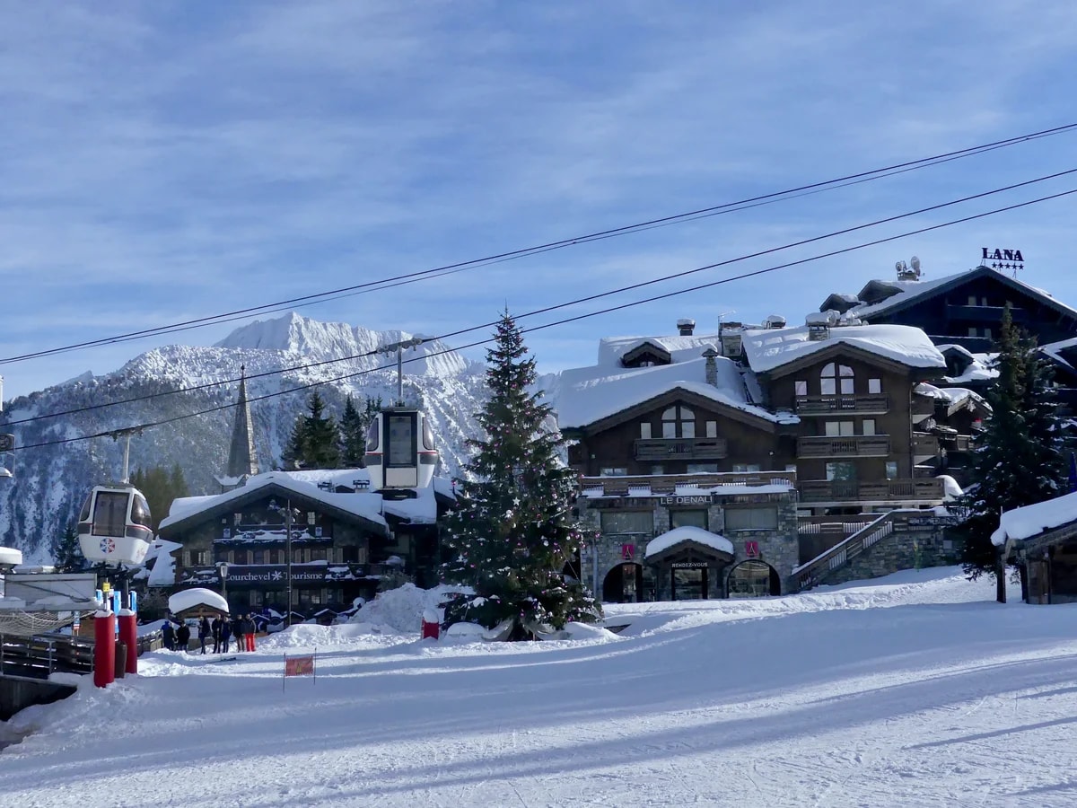 Der Pistenrand von Courchevel 1850 an einem Dezembermorgen, mit verschneiten Chalets, einem Weihnachtsbaum und der Gondelbahn.