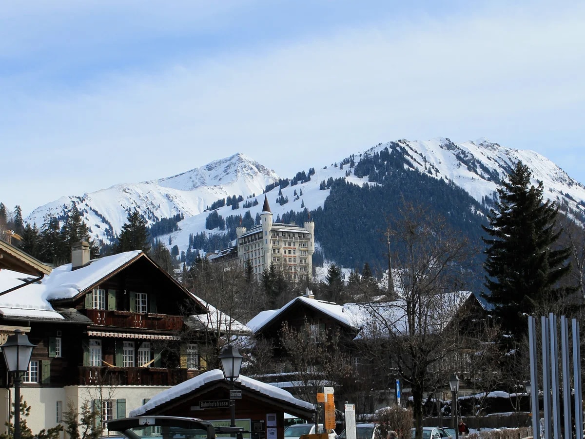 Verschneite Chalets in Gstaad, dahinter ein großes Hotel mit Türmen auf dem Hügel und die Berge.