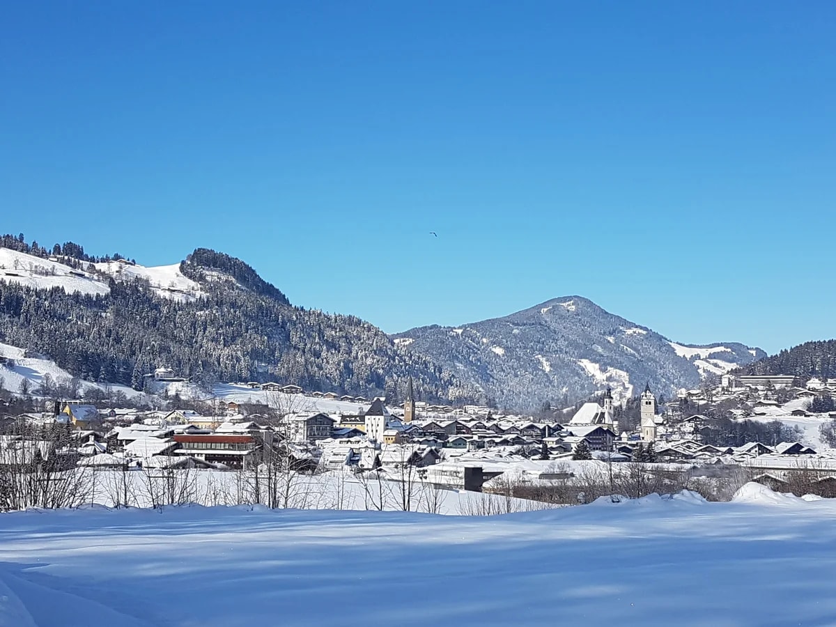 Kitzbühel im Schnee, mit seinen Kirchtürmen am Fuß bewaldeter, verschneiter Berge unter blauem Himmel.