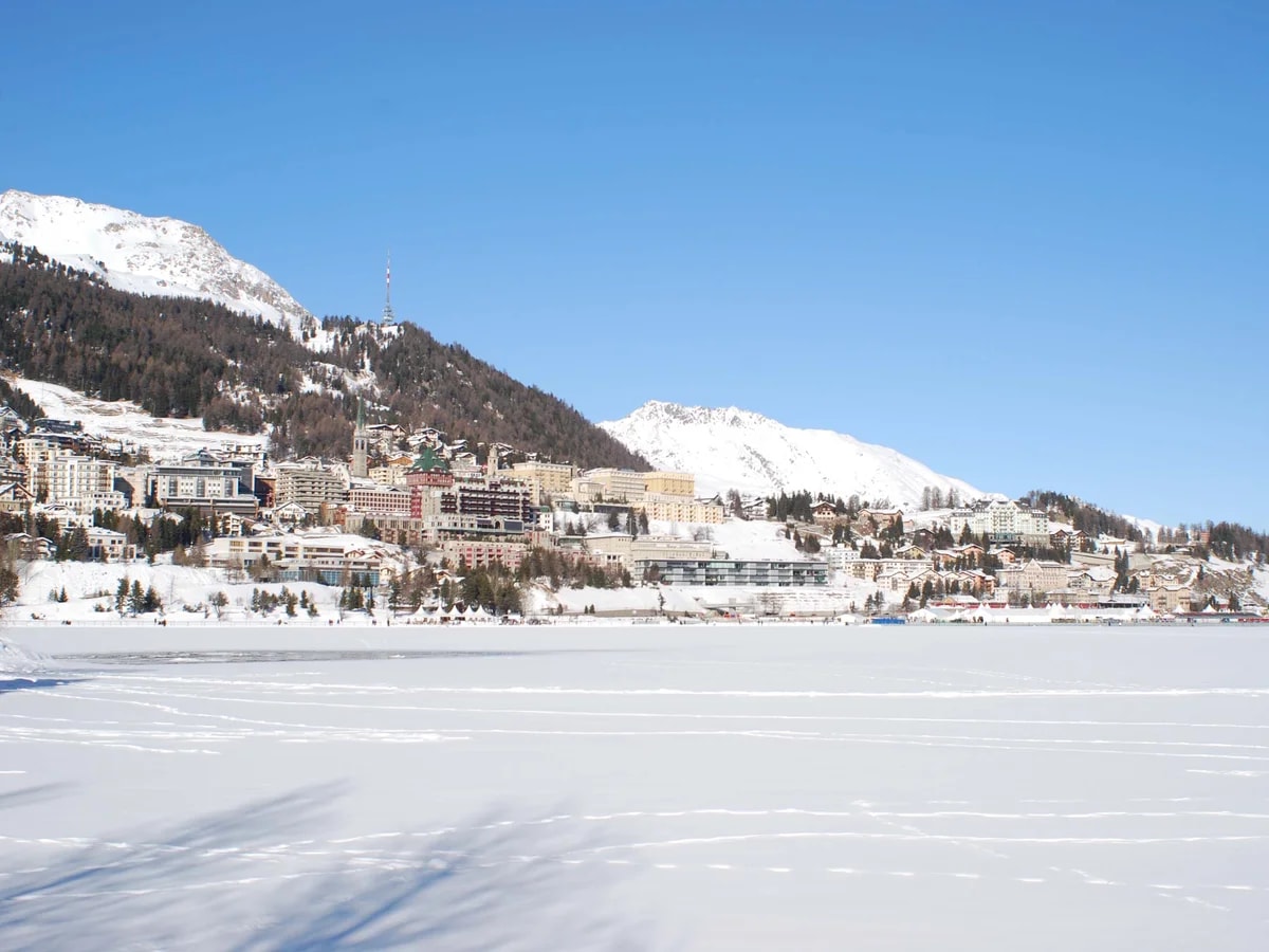 St. Moritz vom zugefrorenen, verschneiten See aus, der Ort am Hang unter den Gipfeln und blauem Himmel.