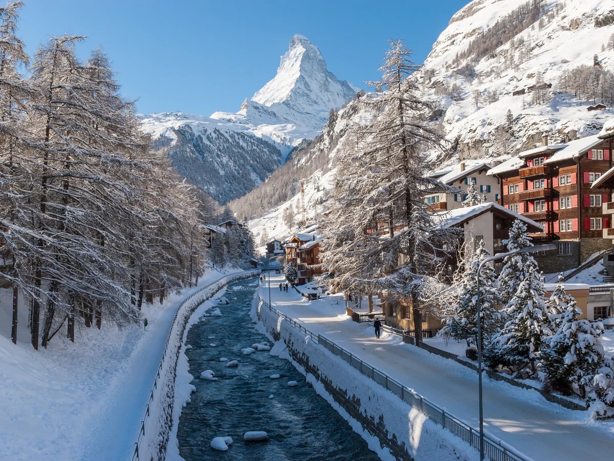 Die Vispa fließt durch das verschneite Zermatt zwischen Holzchalets und Lärchen, dahinter das Matterhorn unter blauem Himmel.